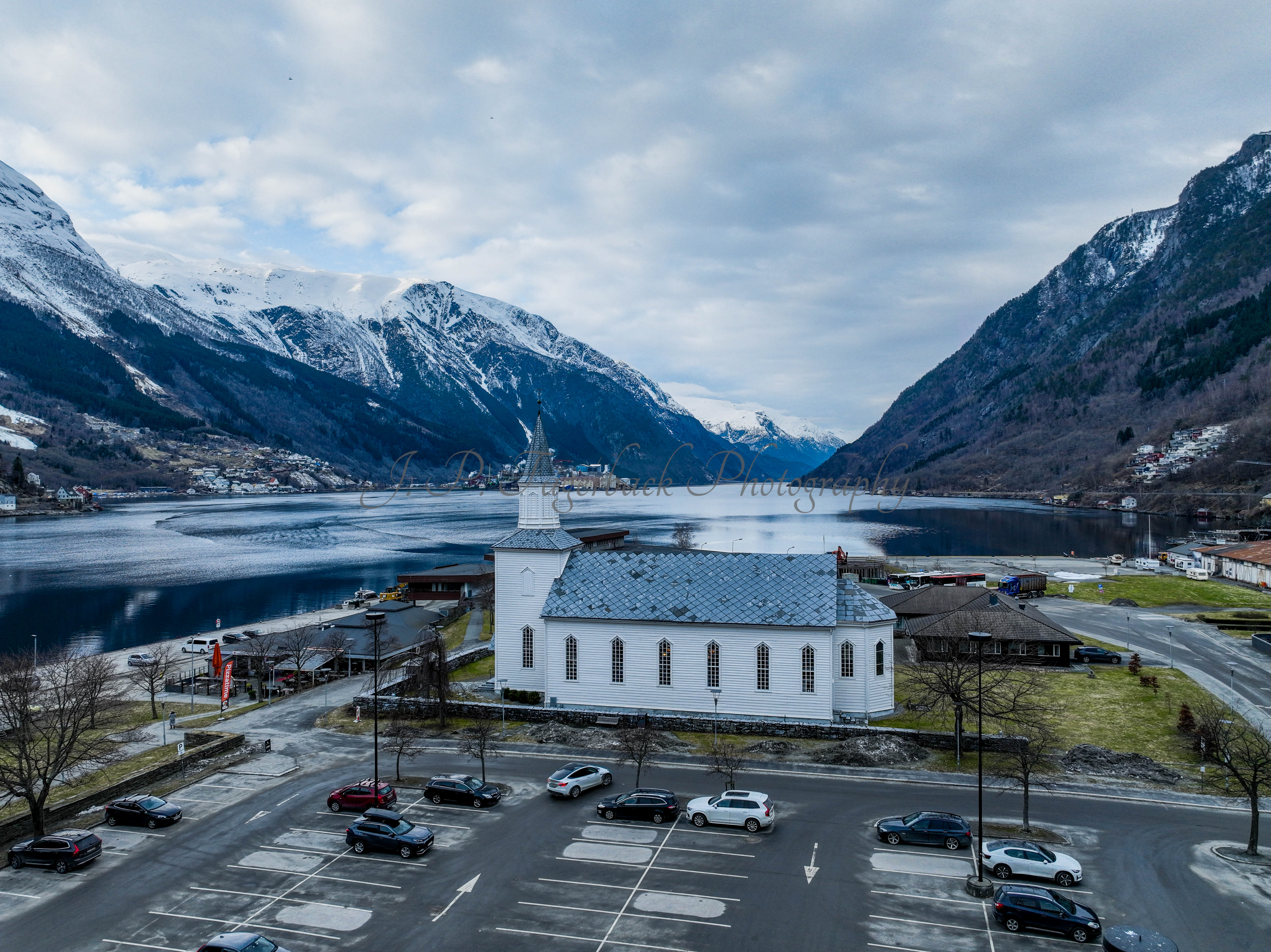 A white church in a town with a parking lot in front and a large body of water behind with land on both sides