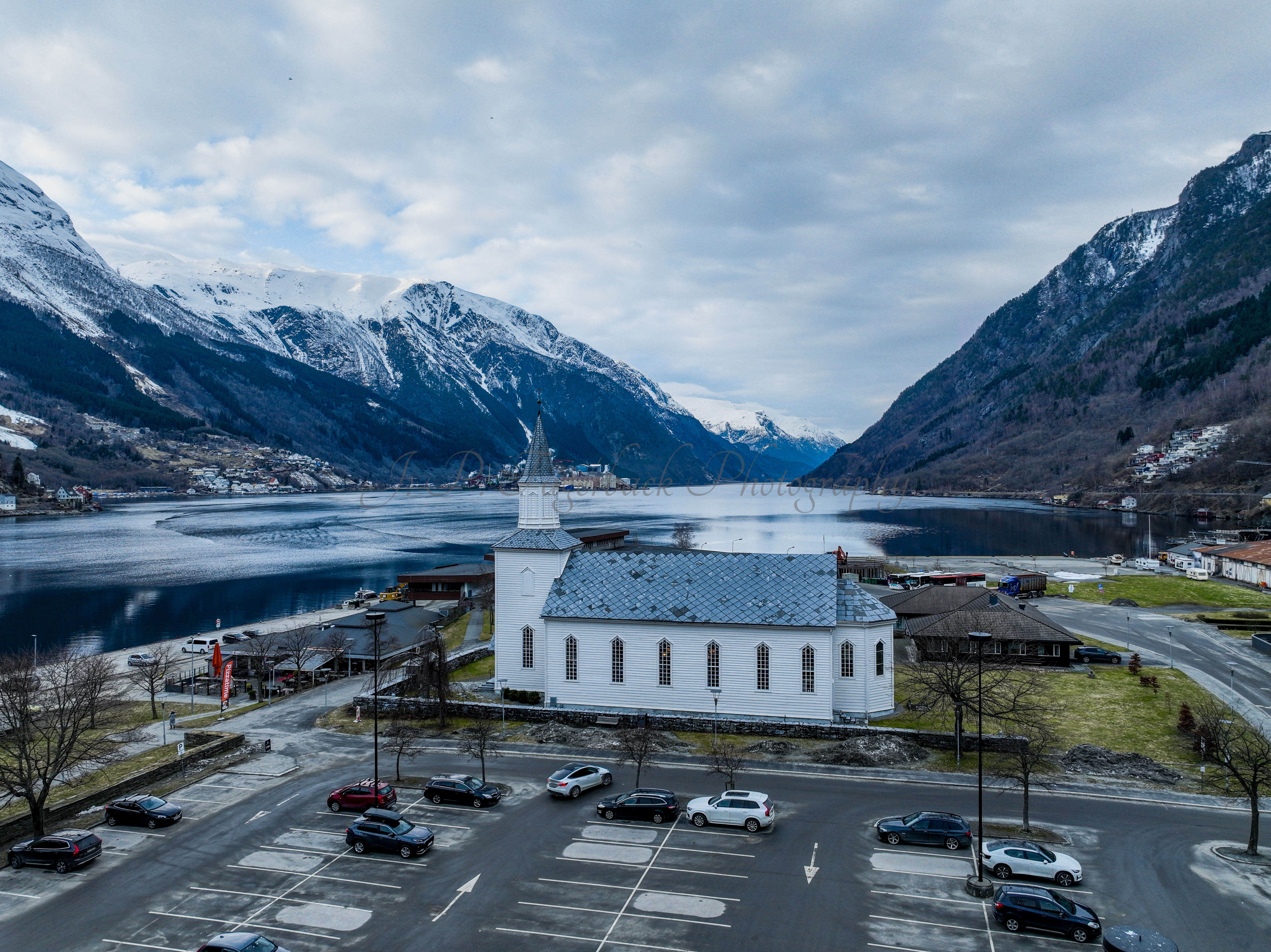 A white church in a town with a parking lot in front and a large body of water behind with land on both sides
