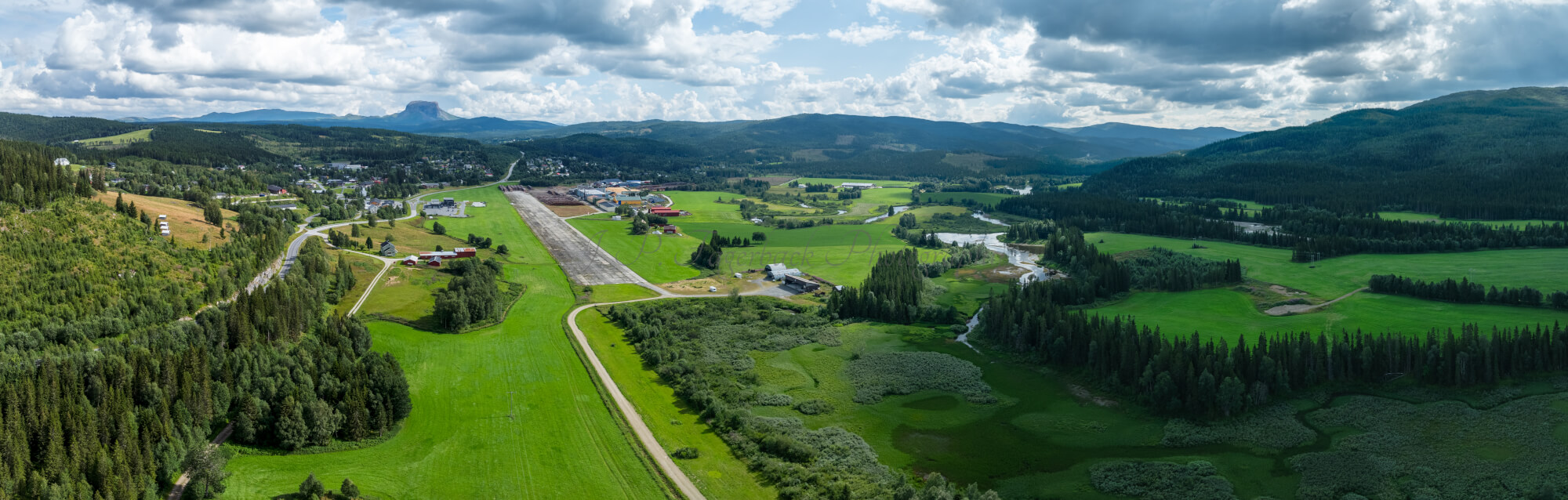 An old airstrip  and fields with buldings beyond it and mountains on the background where one looks like a hat
