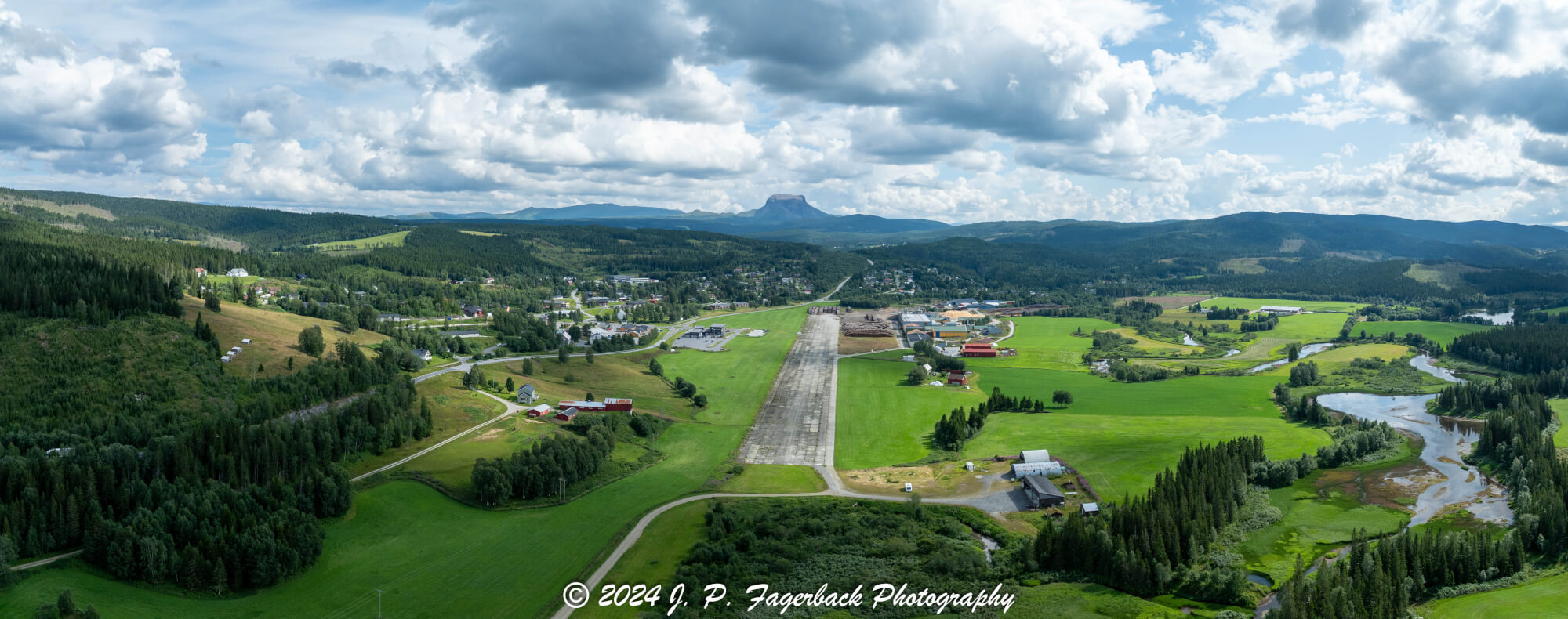 An old airstrip  and fields with buldings beyond it and mountains on the background where one looks like a hat