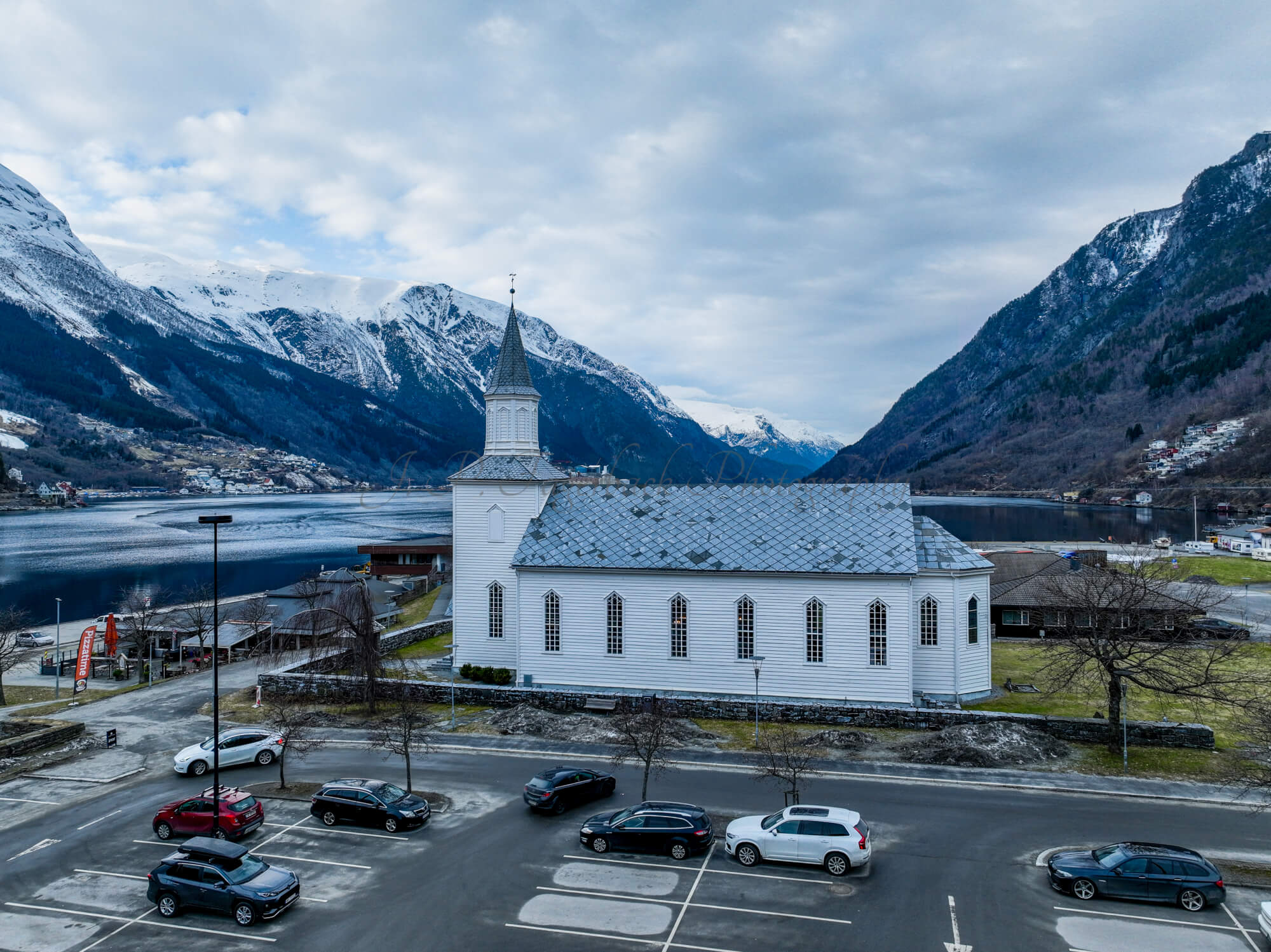 A white church in a town with a parking lot in front and a large body of water behind with land on both sides