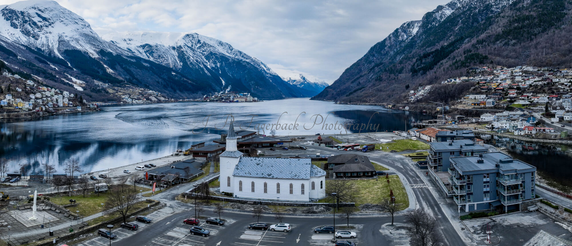 A white church in a town with a parking lot in front and a large body of water behind with land on both sides