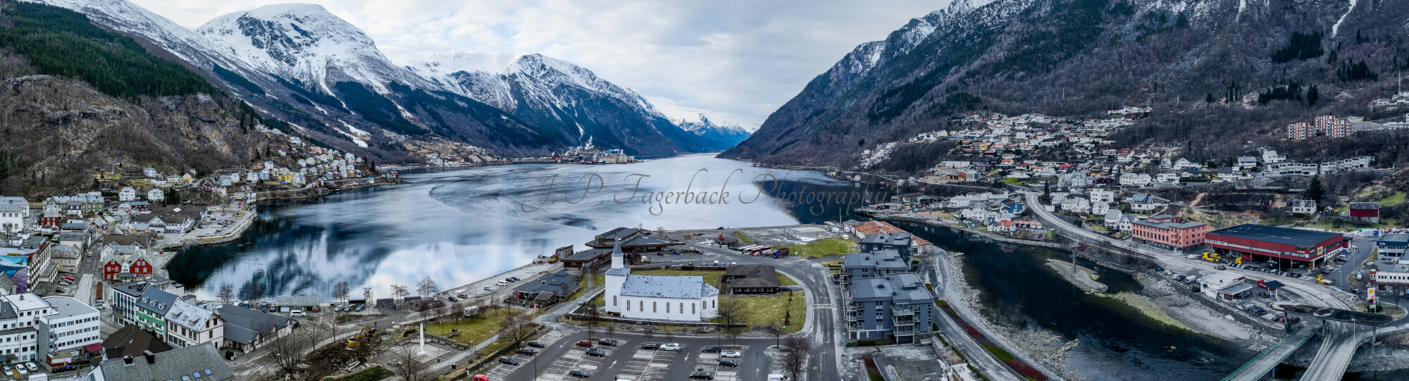 A white church in a town with a parking lot in front and a large body of water behind with land on both sides